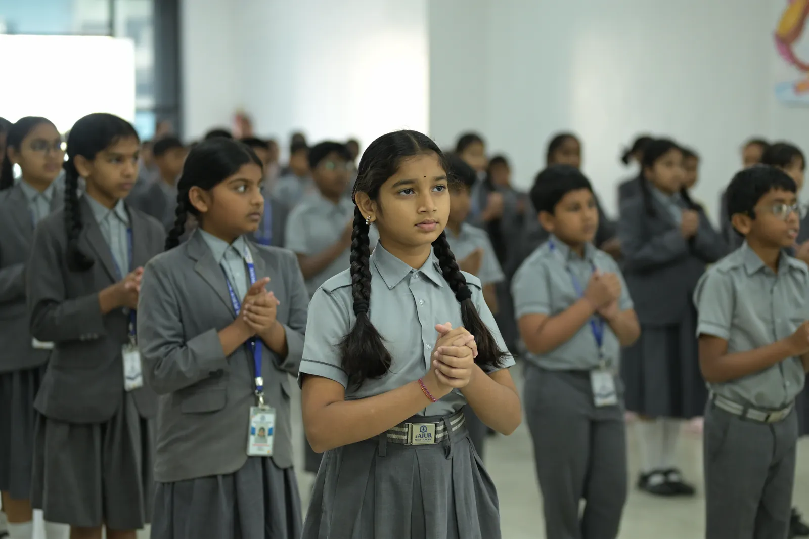 Students standing in rows in the school hall at morning assembly, hands folded.