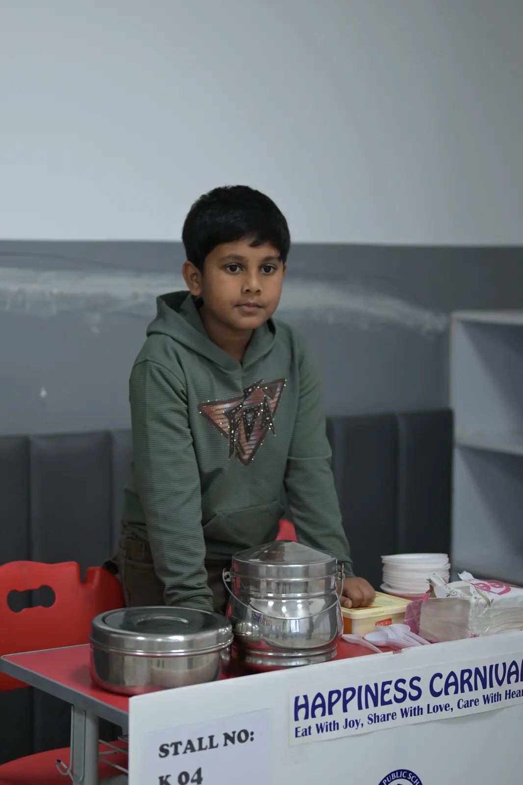 A student minding his stall at the school Happiness Carnival, tiffin containers and serving supplies laid out on the counter in front of him.