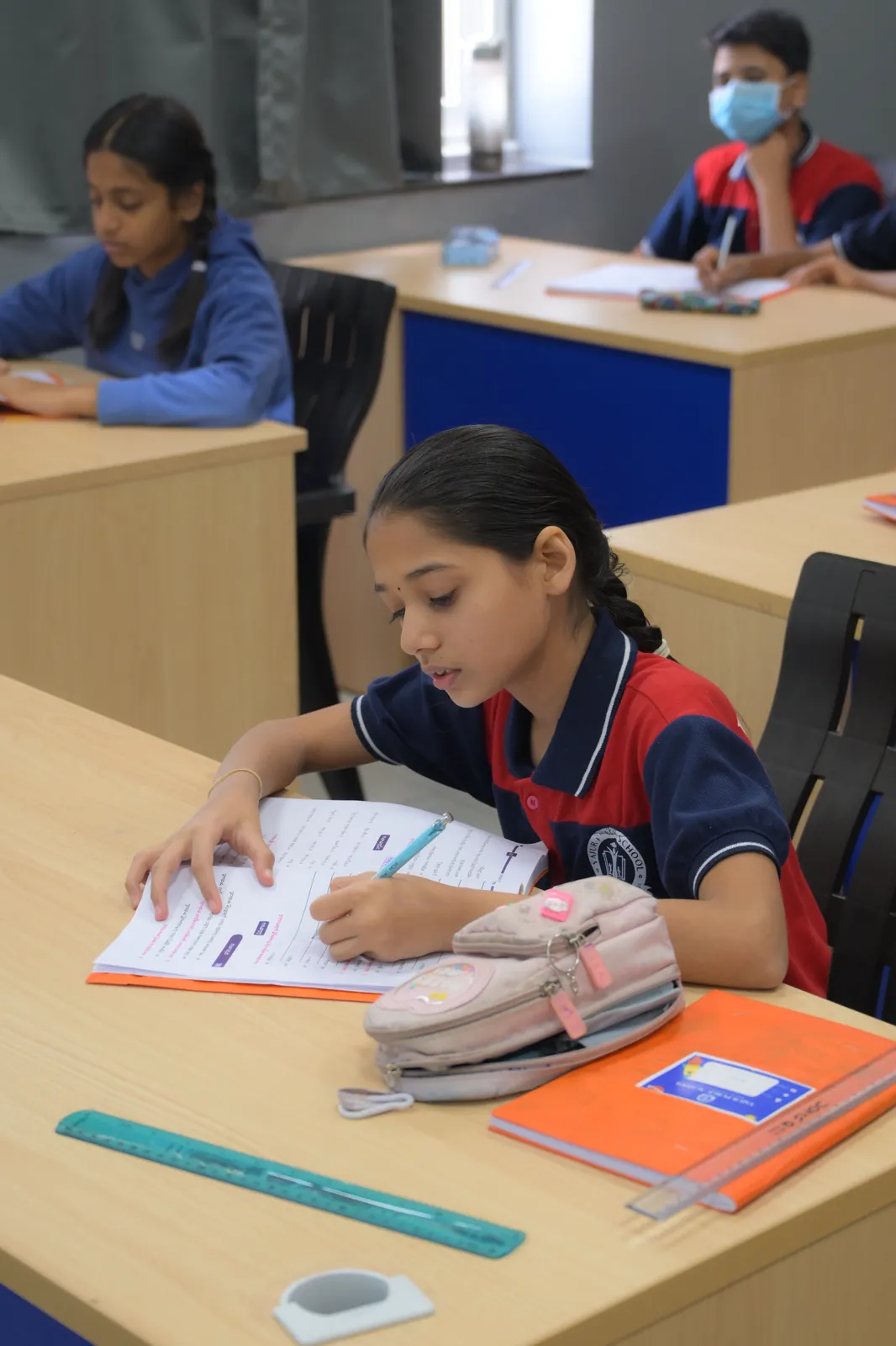 A student writing up a task at her desk, ruler and pencil case laid out beside the book.
