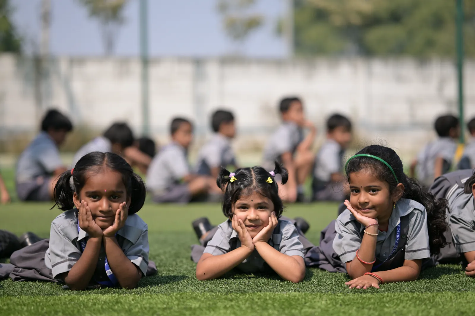 Three students lying on the grass outside, chins propped on their hands, the rest of their class behind them.