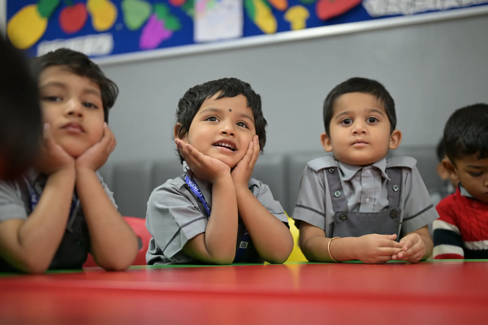 Four of the youngest students at a red classroom table, chins on their hands, following the front of the room.