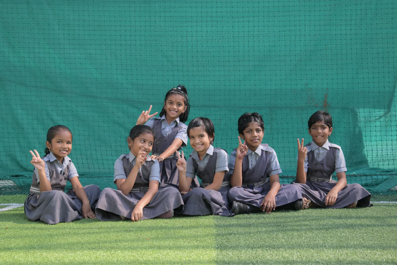 Six students sitting together on the turf at break, holding up hand signs for the camera.